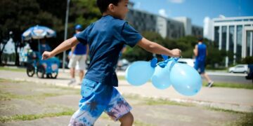 Menino com camiseta azul correndo ao ar livre segurando três balões azuis