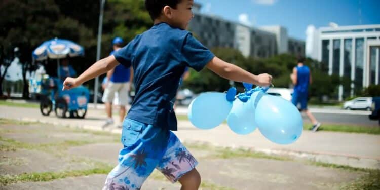 Menino com camiseta azul correndo ao ar livre segurando três balões azuis
