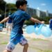 Menino com camiseta azul correndo ao ar livre segurando três balões azuis