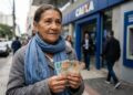 Idosa de cabelos grisalhos segura notas de real em frente à agência da Caixa Econômica Federal