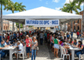 Centenas de pessoas em fila sob tenda branca com faixa "Mutirão do BPC - INSS" em praça de Caruaru com palmeiras e prédios históricos