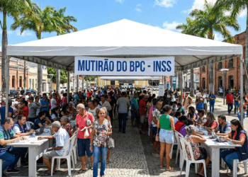 Centenas de pessoas em fila sob tenda branca com faixa "Mutirão do BPC - INSS" em praça de Caruaru com palmeiras e prédios históricos
