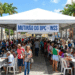 Centenas de pessoas em fila sob tenda branca com faixa "Mutirão do BPC - INSS" em praça de Caruaru com palmeiras e prédios históricos