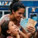 Mulher sorrindo exibe notas de reais com duas crianças em frente à agência Caixa.