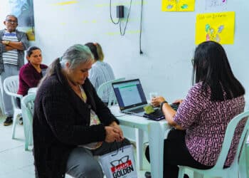 Funcionária do CRAS Bertioga atende mulher idosa em computador, com fila de espera em cadeiras ao fundo