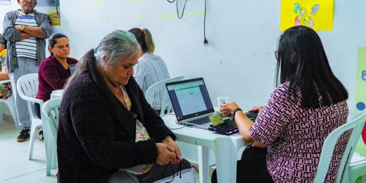 Funcionária do CRAS Bertioga atende mulher idosa em computador, com fila de espera em cadeiras ao fundo