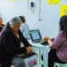 Funcionária do CRAS Bertioga atende mulher idosa em computador, com fila de espera em cadeiras ao fundo