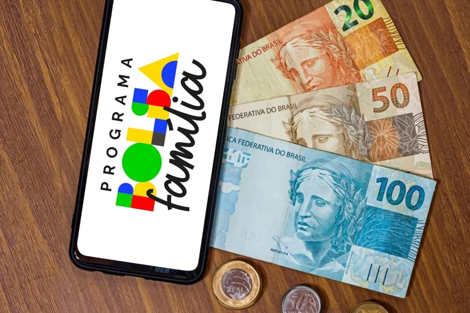Celular com logo do Bolsa Família ao lado de notas de 20, 50 e 100 reais sobre mesa de madeira
