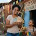 Mulher sorrindo segura dinheiro em notas de real ao lado de duas meninas em festa de São João na rua decorada.