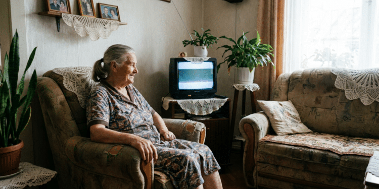 Mulher idosa de cabelos grisalhos sentada em poltrona bege assiste televisão de tubo em sala decorada com crochê e fotos