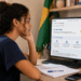 Jovem mulher observa tela de computador com programa Pé-de-Meia do MEC ao lado de caderno e livros
