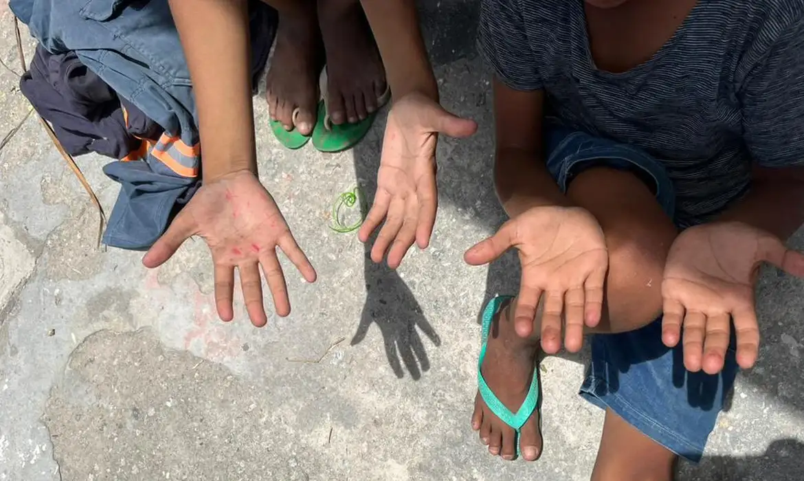 Duas crianças sentadas no chão mostram as mãos abertas enquanto usam chinelos verdes em rua de cimento