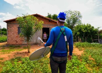 Técnico de uniforme azul e capacete carrega antena parabólica em direção à casa de tijolos em área rural