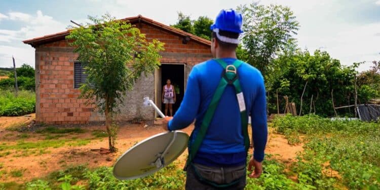 Técnico de uniforme azul e capacete carrega antena parabólica em direção à casa de tijolos em área rural