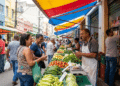 Feirante com avental sorri ao atender clientes em barraca de verduras sob toldo colorido em feira livre urbana