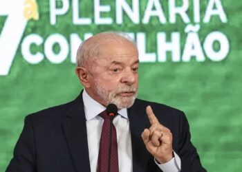 Lula em paletó escuro e gravata, fala ao microfone, dedo em riste, painel verde com “7ª Plenária Conselhão” ao fundo