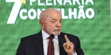 Lula em paletó escuro e gravata, fala ao microfone, dedo em riste, painel verde com “7ª Plenária Conselhão” ao fundo
