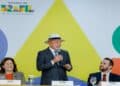Lula de chapéu e terno discursa sorrindo entre ministros em mesa com logomarca do governo Brasil