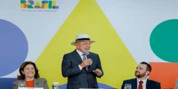 Lula de chapéu e terno discursa sorrindo entre ministros em mesa com logomarca do governo Brasil