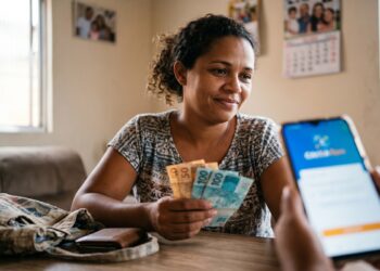 Mulher sorrindo segura notas de 50 e 100 reais enquanto outra pessoa mostra o aplicativo Caixa Tem no celular
