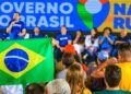 Pessoas assistem a evento do Governo do Brasil, com painel azul, bandeira nacional e participantes no palco