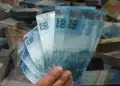 Mão segurando notas de 100 reais com pilhas de dinheiro ao fundo.