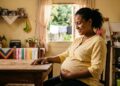 Mulher grávida sentada à mesa, sorrindo e segurando a barriga em cozinha simples iluminada