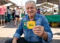 Homem idoso sentado ao ar livre sorrindo e exibindo Carteira do Idoso em feira