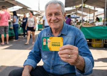 Homem idoso sentado ao ar livre sorrindo e exibindo Carteira do Idoso em feira