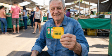 Homem idoso sentado ao ar livre sorrindo e exibindo Carteira do Idoso em feira