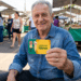 Homem idoso sentado ao ar livre sorrindo e exibindo Carteira do Idoso em feira