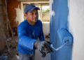 Pintor com boné e luvas aplica tinta azul em parede com rolo em obra residencial