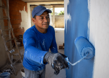 Pintor com boné e luvas aplica tinta azul em parede com rolo em obra residencial