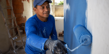 Pintor com boné e luvas aplica tinta azul em parede com rolo em obra residencial