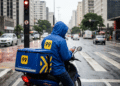 Entregador da 99 com capa de chuva azul sobre moto verifica celular em semáforo durante dia chuvoso em cidade brasileira