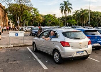 Homem idoso em Chevrolet Onix prata estaciona em vaga exclusiva para idosos com placa "ESTACIONAMENTO - IDOSOS"