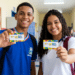 Jovens estudantes sorrindo exibem cartões do Pé-de-Meia em corredor de escola, com uniformes azul e branco