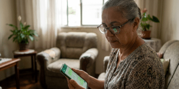 Mulher idosa de cabelos grisalhos e óculos lê mensagem no smartphone em sala de estar decorada com plantas