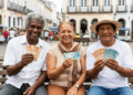 Três idosos sorridentes exibem cédulas de real em banco de madeira em praça colonial brasileira