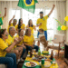 Família com camisas amarelas, pintura facial verde-amarela, bandeira do Brasil, celebrando gol em TV com placar Brasil 2x0 Argentina