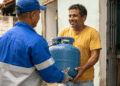 Homem sorridente recebe botijão de gás azul de entregador uniformizado na porta de casa