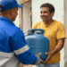 Homem sorridente recebe botijão de gás azul de entregador uniformizado na porta de casa
