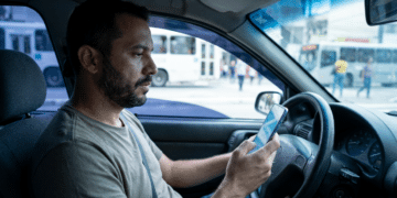Homem barbudo de camiseta cinza segura celular com mapa ao volante, ônibus urbanos desfocados ao fundo