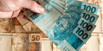 Mão segurando várias cédulas de 100 reais sobre notas de 50 reais brasileiras.