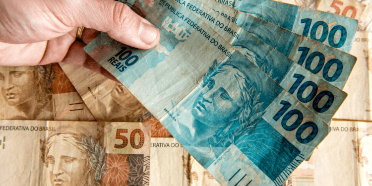 Mão segurando várias cédulas de 100 reais sobre notas de 50 reais brasileiras.