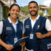 Dois agentes de saúde do SUS sorrindo em rua residencial, usando coletes azuis e crachás