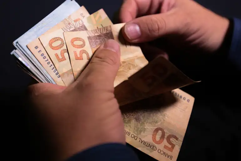 Mãos masculinas contando várias notas de 50 reais em ambiente escuro