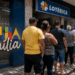 Pessoas em fila na porta de uma lotérica ao lado do letreiro Bolsa Família em ambiente urbano