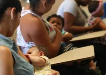 Quatro mulheres amamentam seus bebês sentadas em carteiras escolares, ambiente de sala de aula