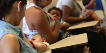 Quatro mulheres amamentam seus bebês sentadas em carteiras escolares, ambiente de sala de aula
